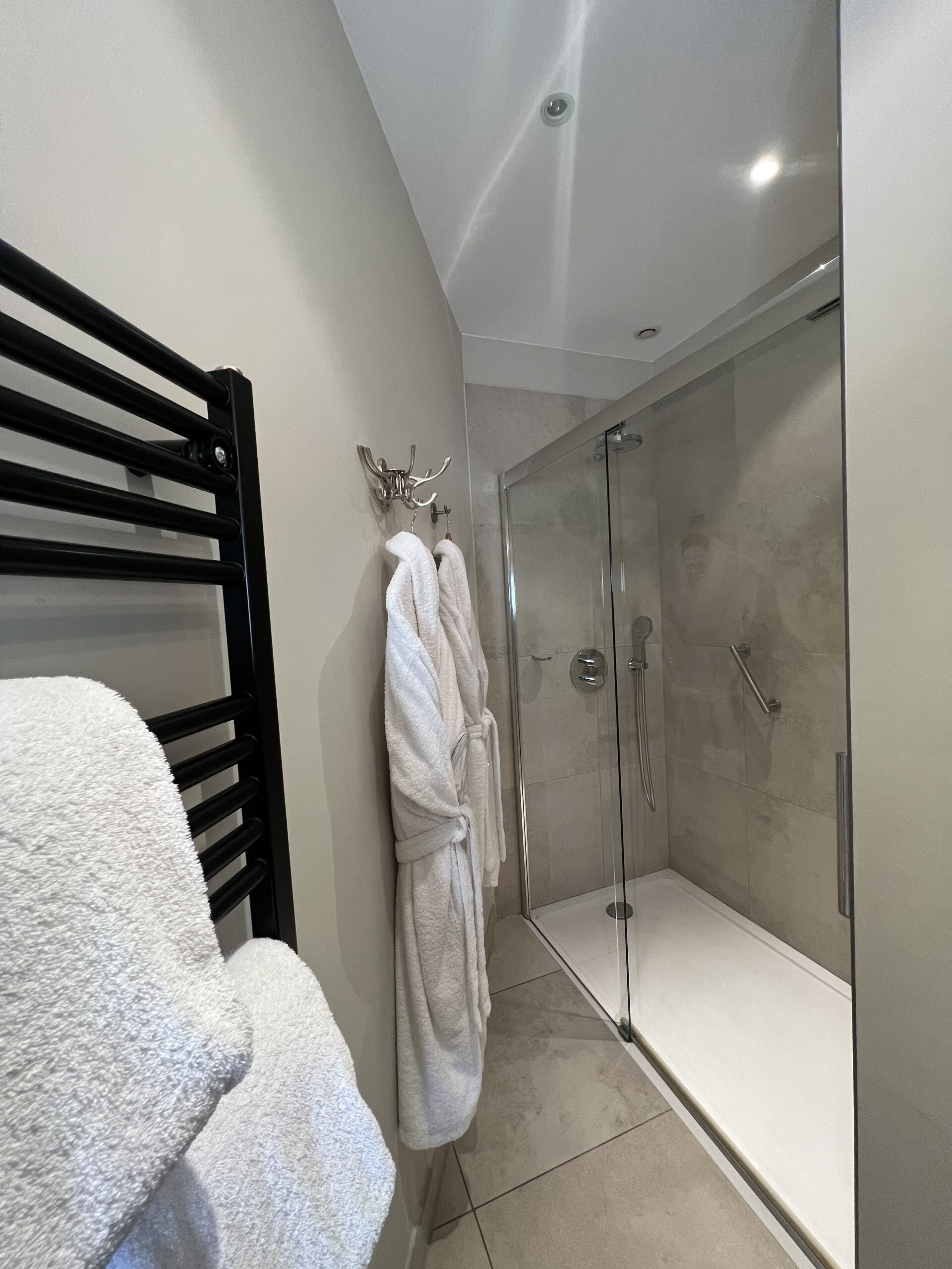 Un baño con ducha envuelta en vidrio, un perchero negro y una bata blanca colgada en la pared.