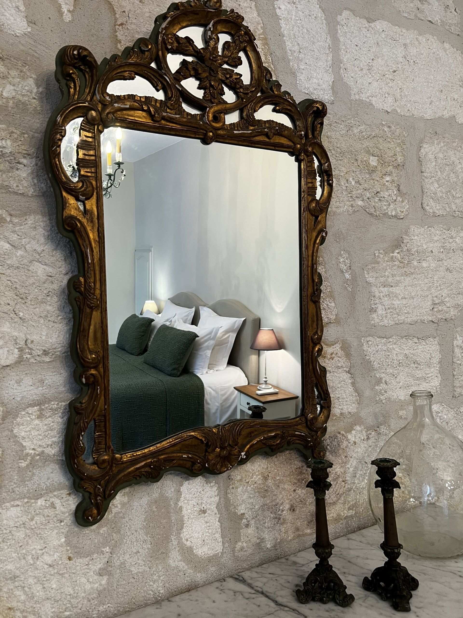 A mirror with an ornate frame reflecting a bedroom with a bed, pillows, a nightstand, and a lamp.