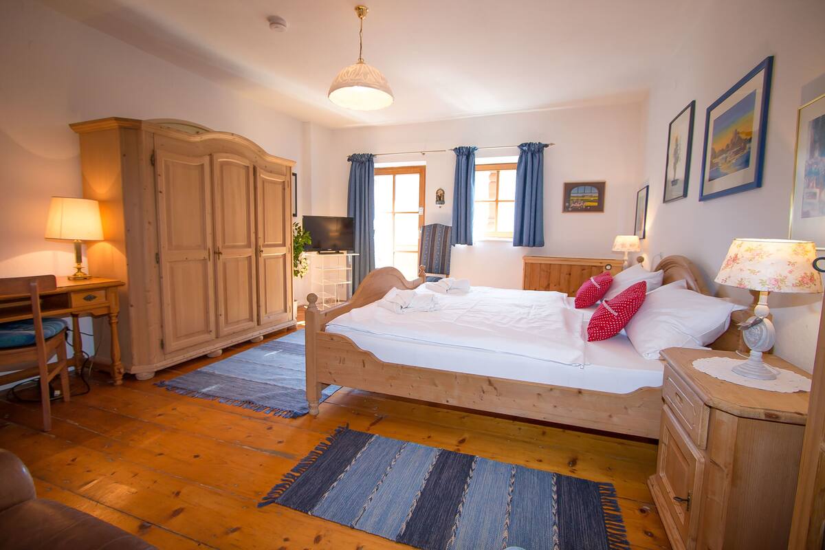 A bedroom with a wooden bed, a desk with a lamp, a wardrobe, and a television on a stand.
