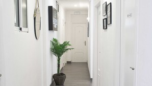 A hallway with white walls, a potted plant, framed pictures, and a door at the end. - Hostal Guestped (Madrid)