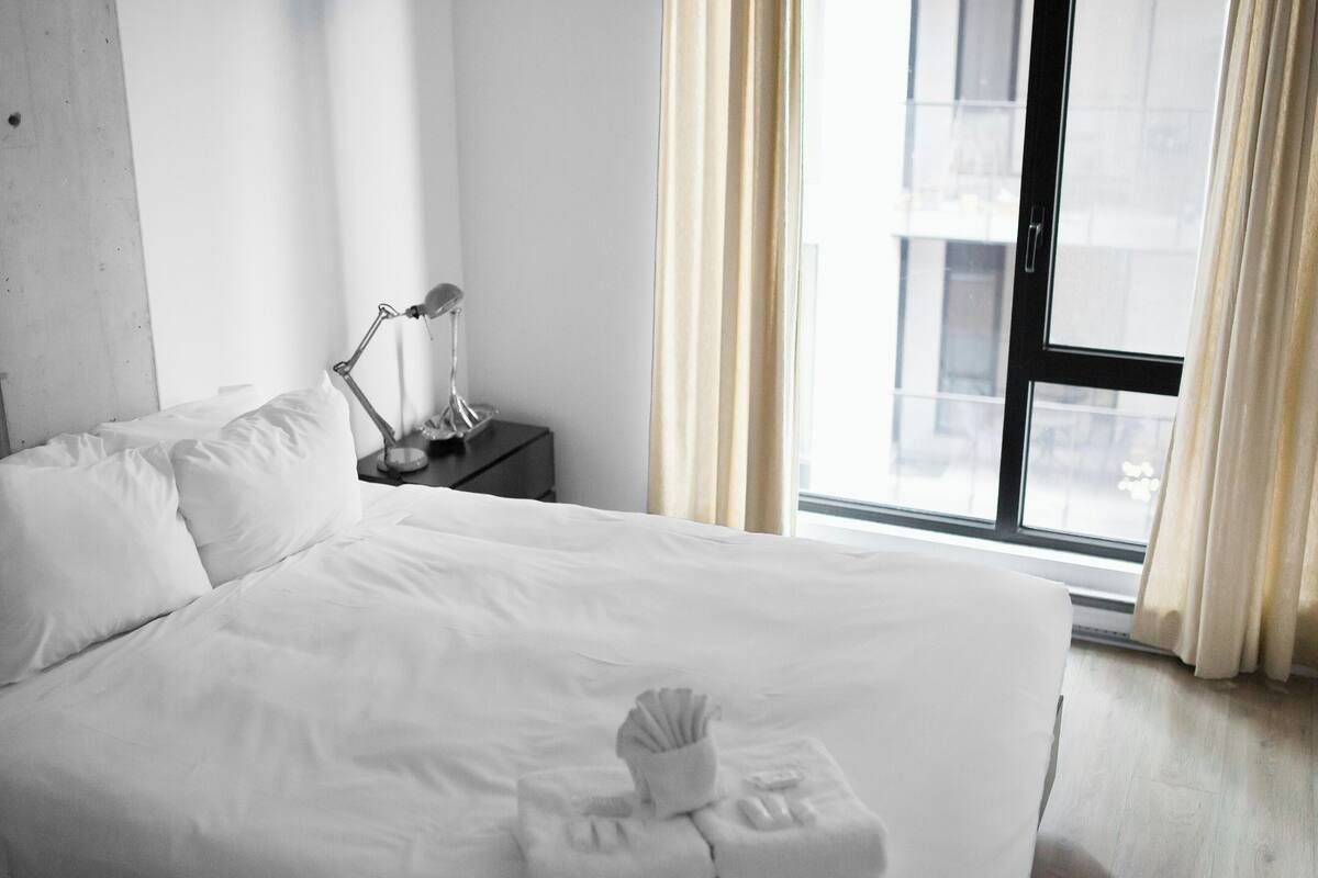 A hotel room with a large bed, a bedside table with a lamp, and a window with curtains.