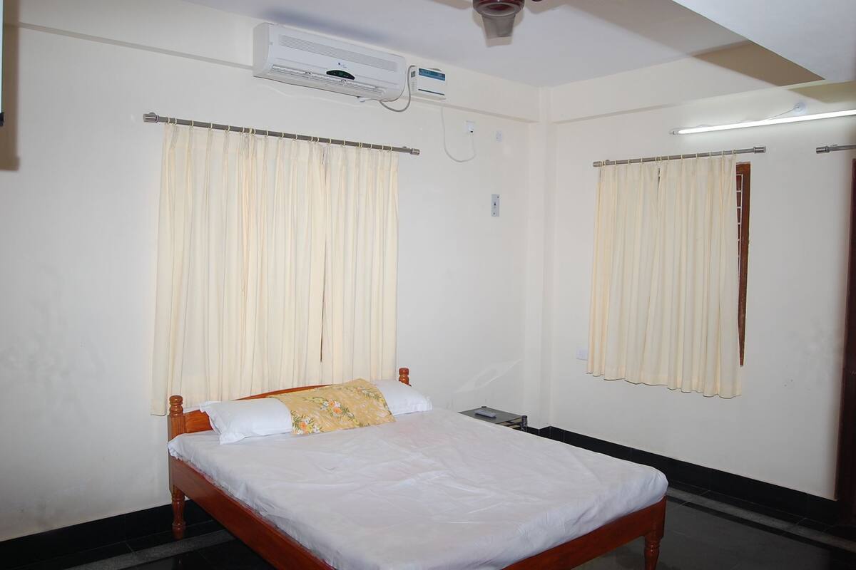A single bed with a wooden frame, a white mattress, and a floral pillow. There is a ceiling fan, an air conditioning unit, and a mirror on the wall.