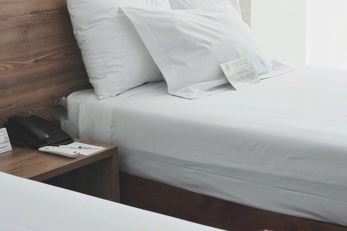 A neatly made bed with white linens, a wooden headboard, and a bedside table with a remote control and a phone.