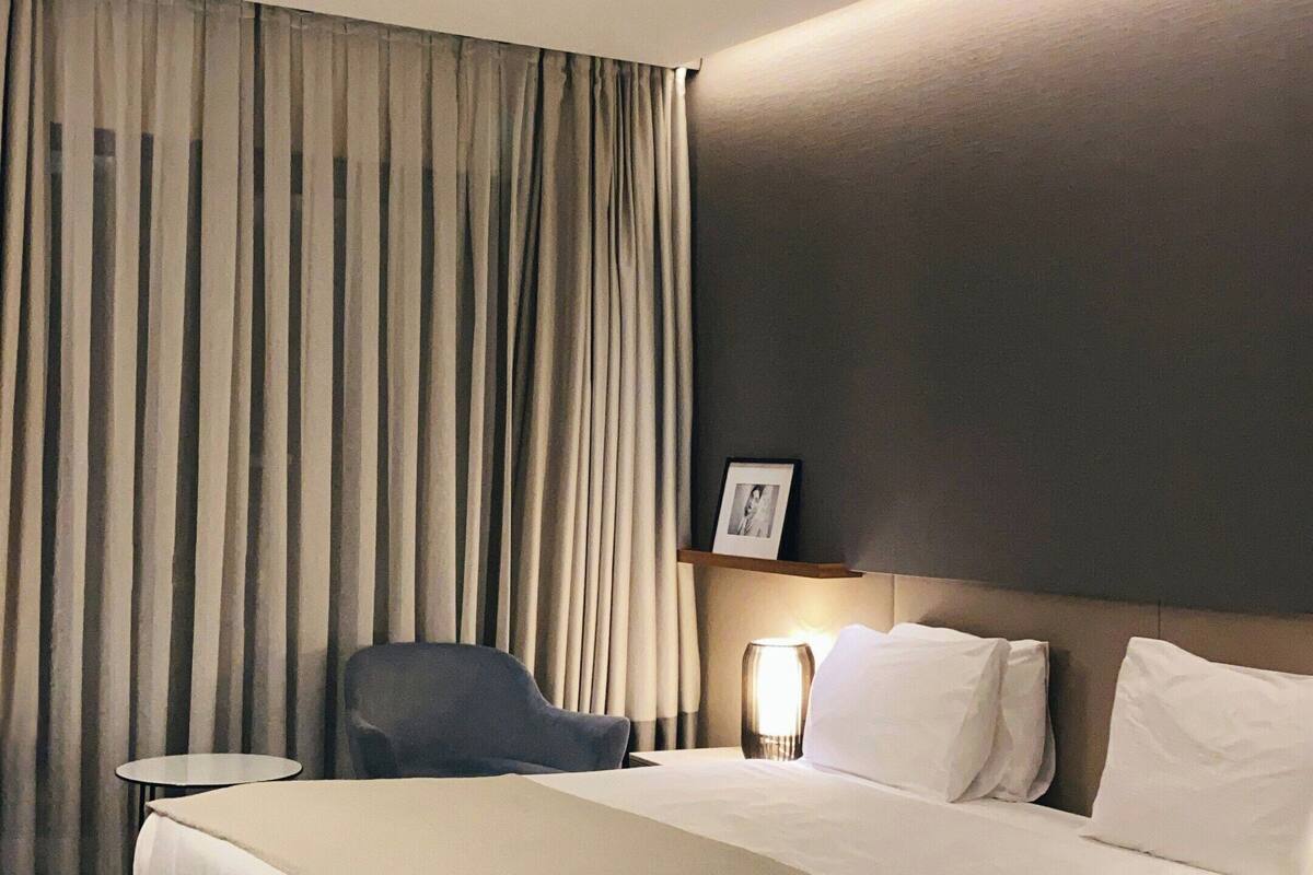 A hotel room with a large bed, a bedside table, a chair, a lamp, and a curtain.