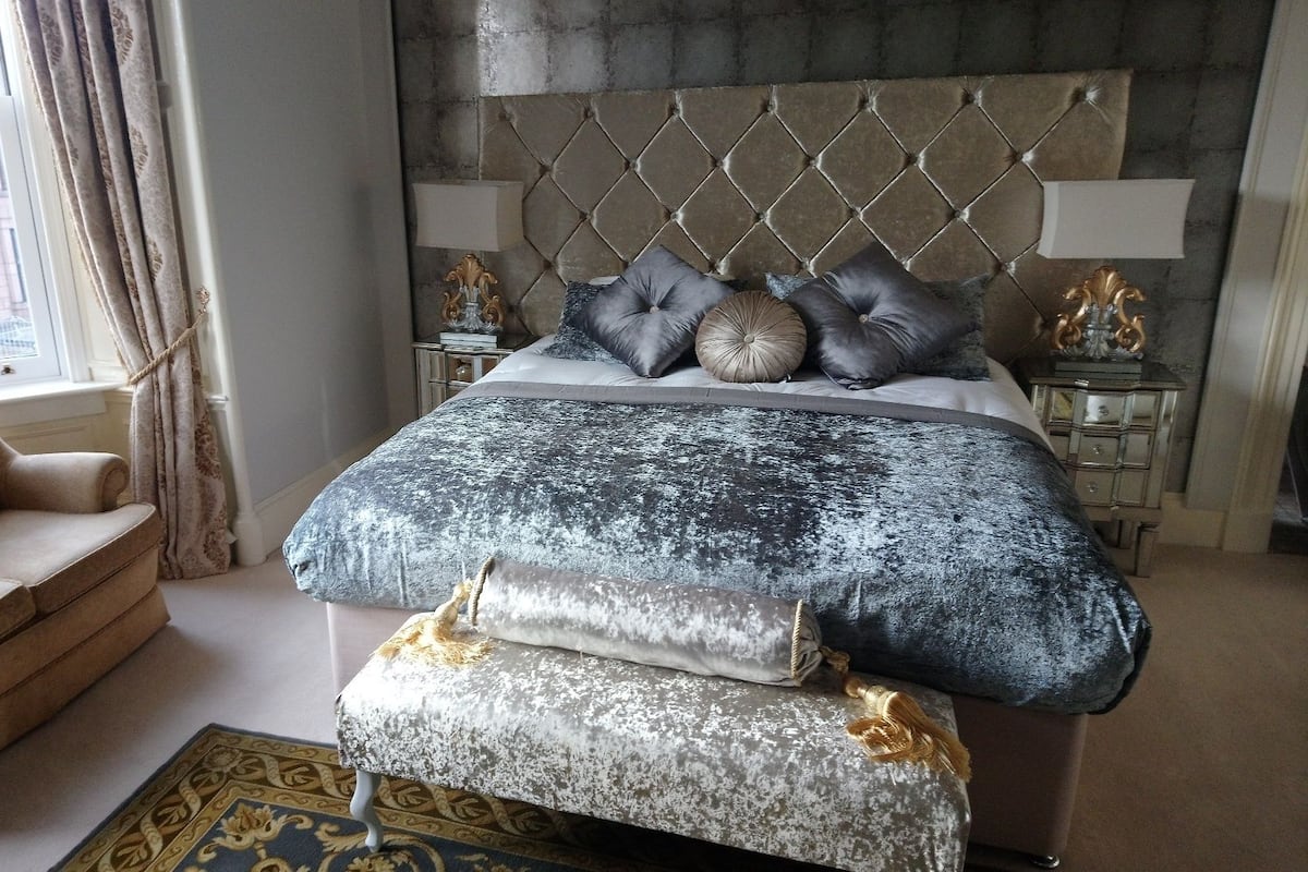 A bedroom with a tufted headboard, a large bed with multiple pillows, a chaise lounge, and a decorative lamp on a nightstand.