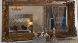Une chambre à coucher avec un grand miroir décoratif, un lustre, un lit doté d’une tête de lit capitonnée et plusieurs carafes en cristal disposées sur une surface en marbre.