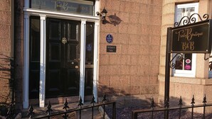 Une porte noire à deux battants avec une poignée en laiton, une enseigne au-dessus indiquant « Trinity Boutique B&B », et une plaque indiquant l’adresse « 104, rue Queen, Trinity ».