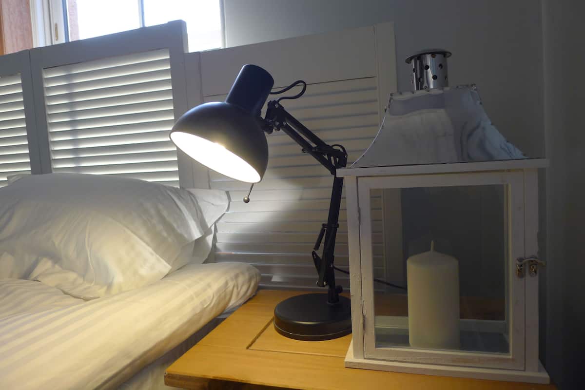 A bedside table with a lamp, a white candle in a glass container, and a window with blinds.