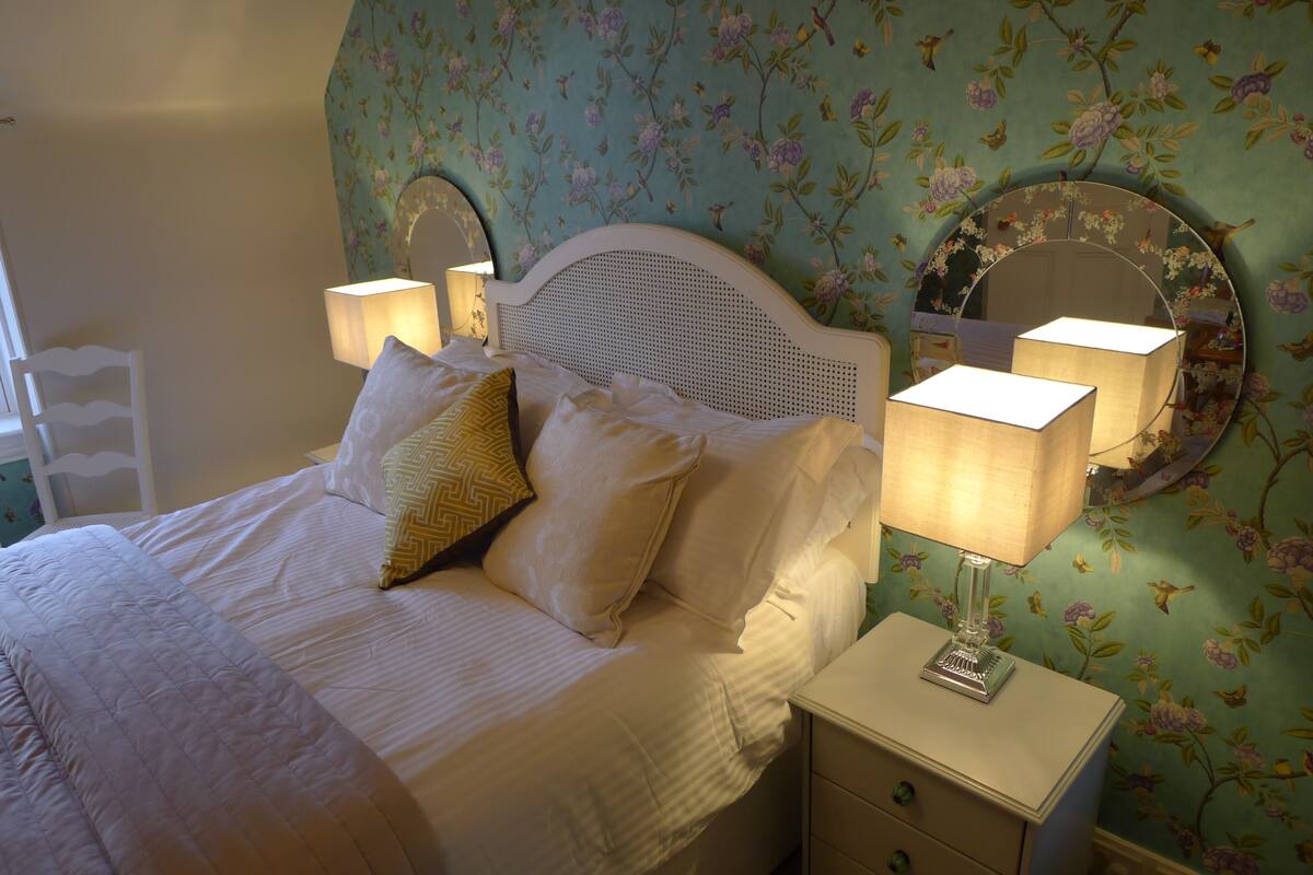 A neatly made bed with white linens and decorative pillows, a bedside table with a lamp, and floral wallpaper.