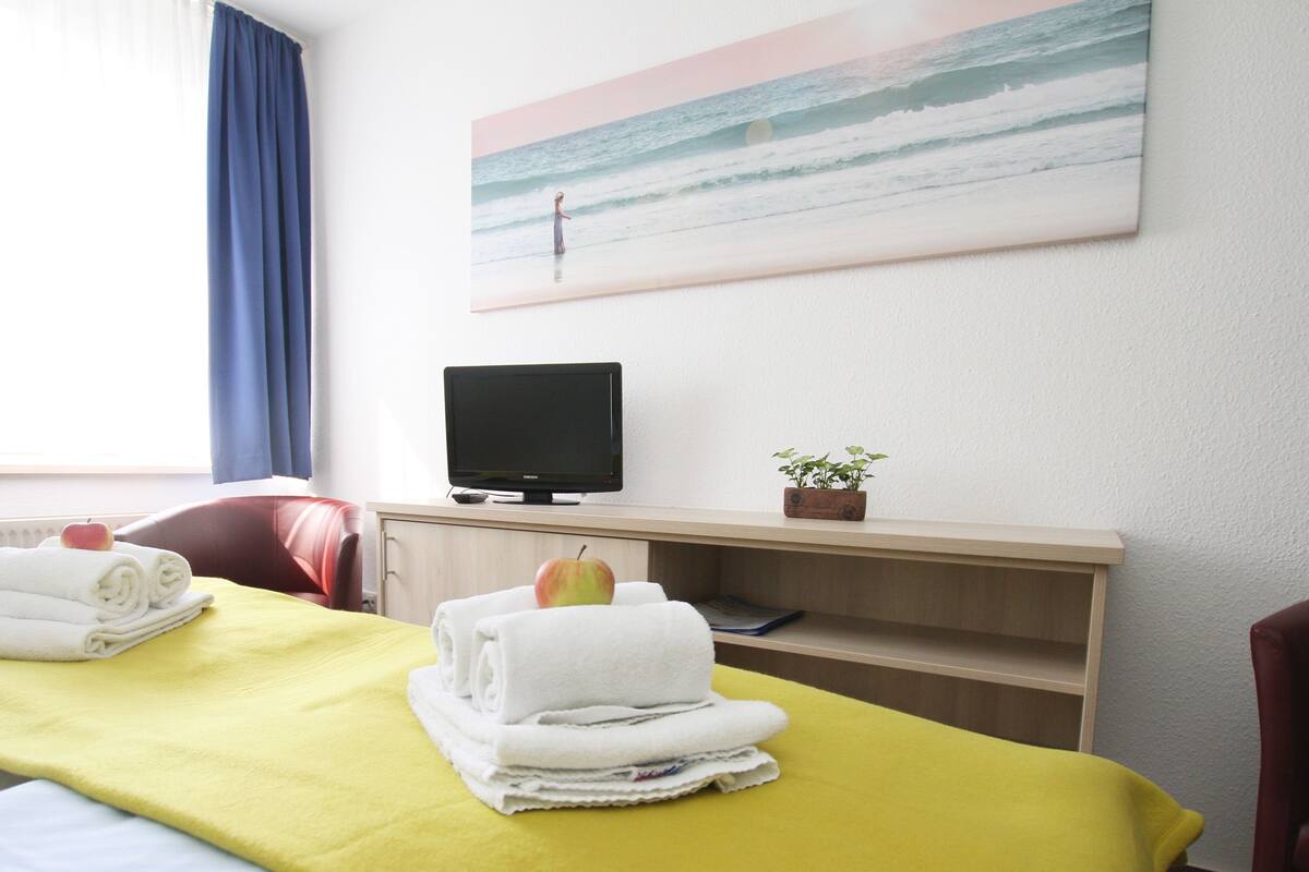 A hotel room with a bed, a desk with a computer, a TV, and a painting of a beach scene.