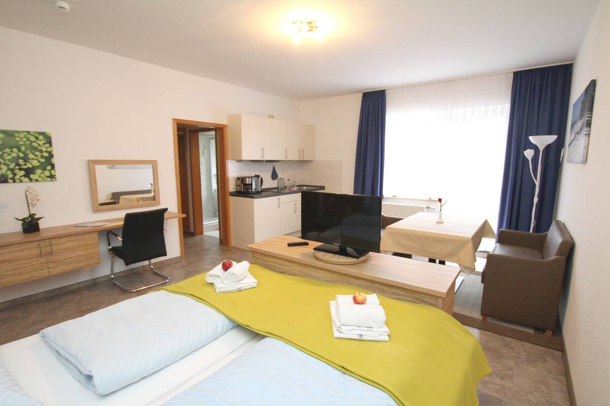 A hotel room with a bed, a desk with a computer, a television, a chair, and a small kitchenette.