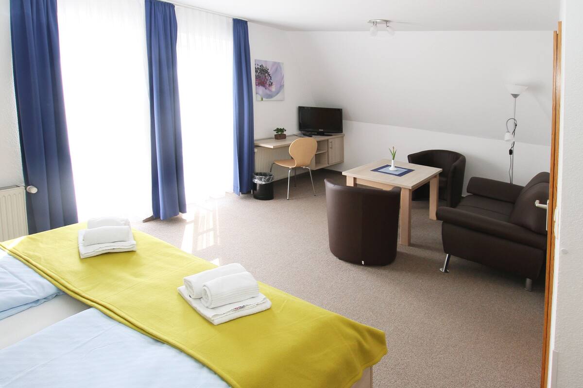 A hotel room with two beds, a desk, a chair, a sofa, a TV, and a lamp.