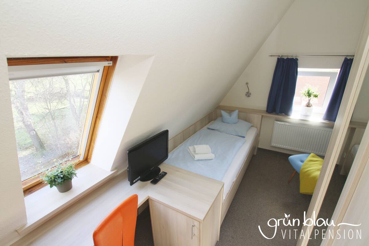 A small, tidy room with a bed, a desk with a computer, a chair, and a window with a plant.