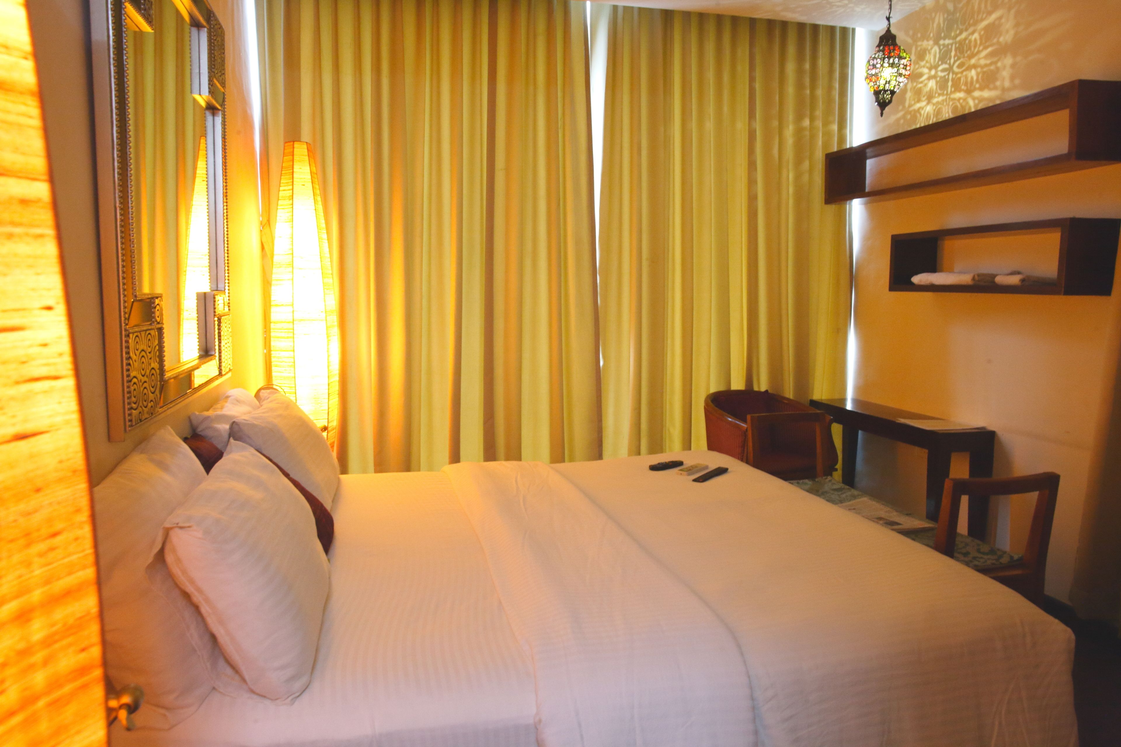 A hotel room with a bed, a bedside table, a mirror, a shelf with towels, and a window with curtains.