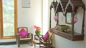 Una habitación con dos sillas de madera, un jarrón verde con flores, un espejo con marco decorativo y una ventana que da a unas árboles.