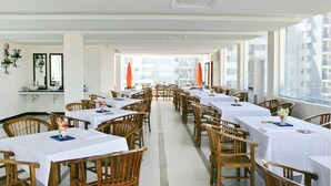 Un restaurante con varias mesas y sillas, amplias ventanas con vistas a edificios y un interior en tonos blancos.