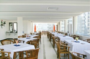 Un restaurante con varias mesas y sillas, amplias ventanas con vistas a edificios y un interior en tonos blancos.
