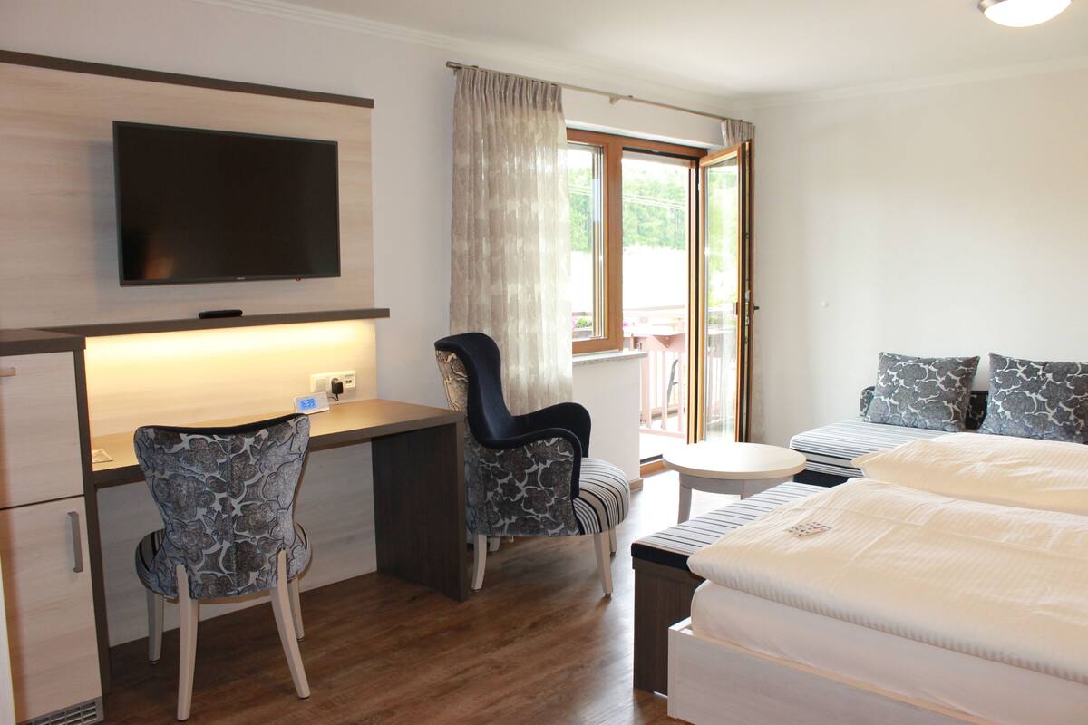 Et hotelværelse med en seng, to stole, et skrivebord, et fjernsyn og et vindue med gardiner.