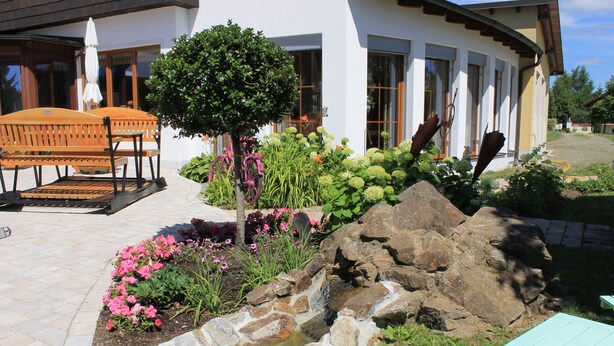 A patio with outdoor seating, a small water feature, and a garden with various plants and flowers. - Landhotel Kastanienhof (Georgenberg)