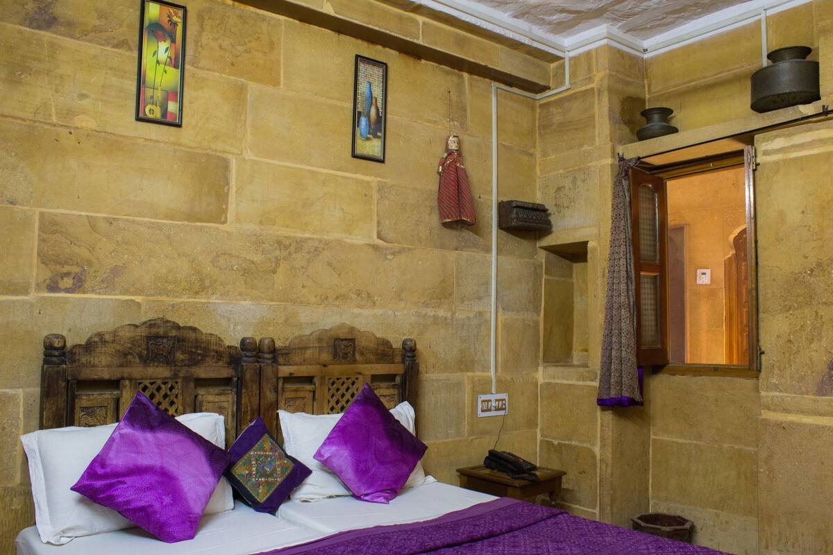 A room with stone walls, a wooden bed with purple pillows, a small table, and a window with a curtain.