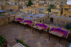A rooftop terrace with multiple seating areas, each with a table and cushions. The terrace is surrounded by traditional buildings with flat roofs and balconies. - Hotel Shahi Palace (Jaisalmer)