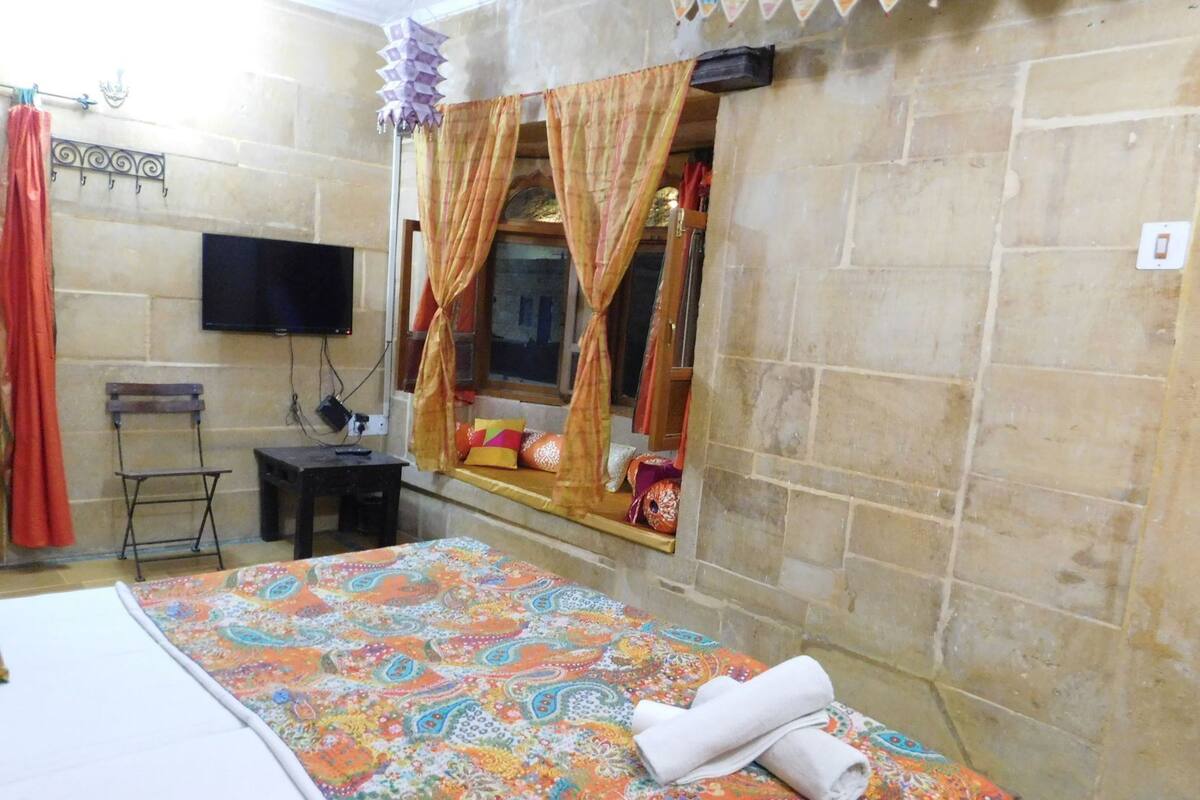 A room with a bed, a small table, a chair, a television mounted on the wall, and a window with curtains.