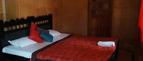 Una cama con cabecera de madera, colcha estampada roja y almohadas en rojo, azul y blanco.