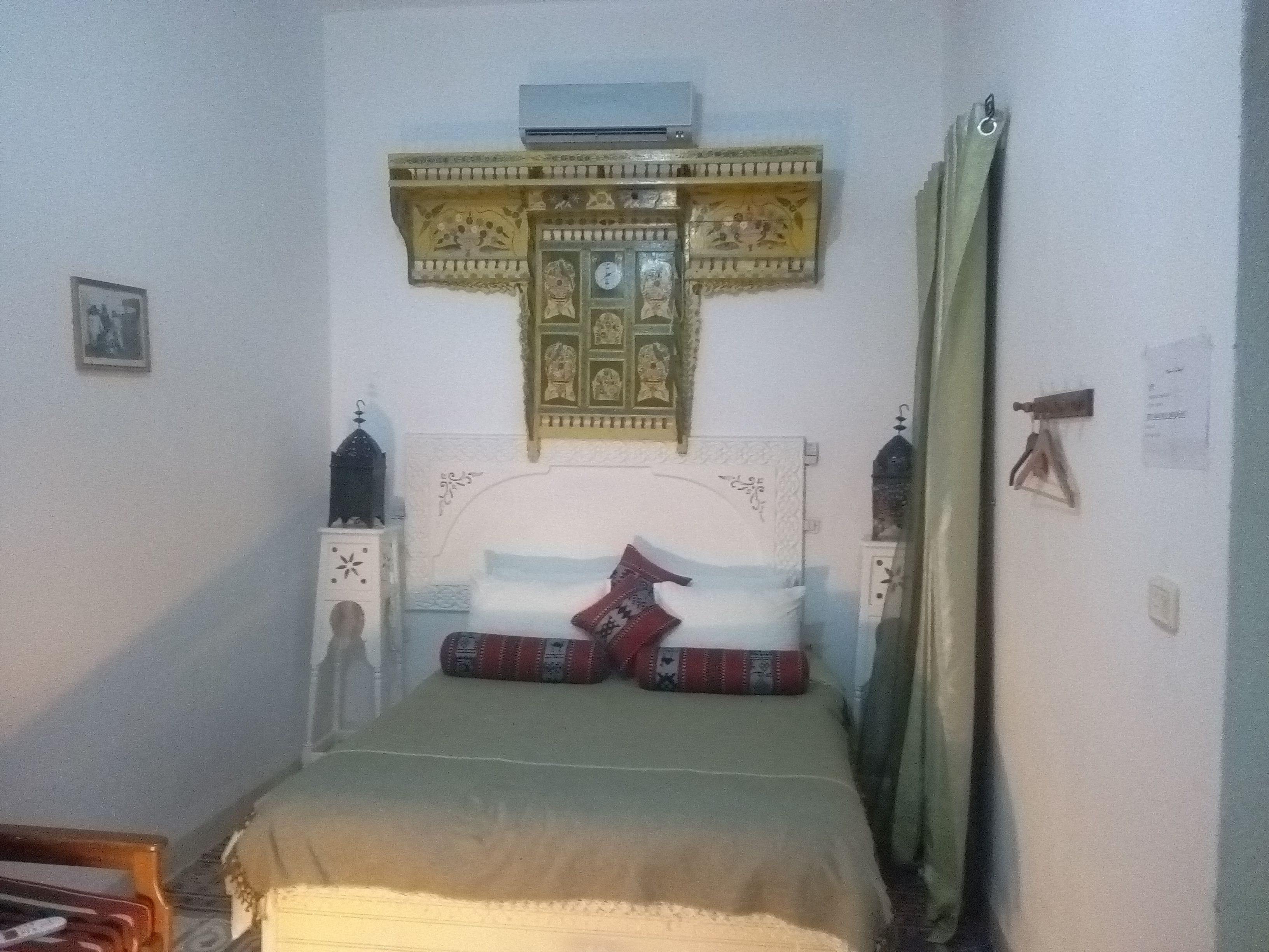 A bedroom with a bed, a wall-mounted air conditioner, a decorative cross, and a small cabinet.