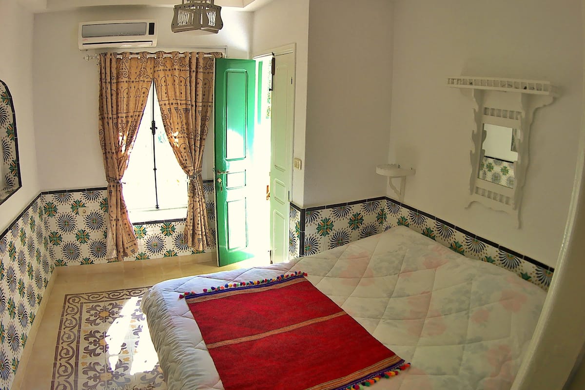 A bedroom with a bed, a green door, patterned walls, a hanging light fixture, and a window with curtains.