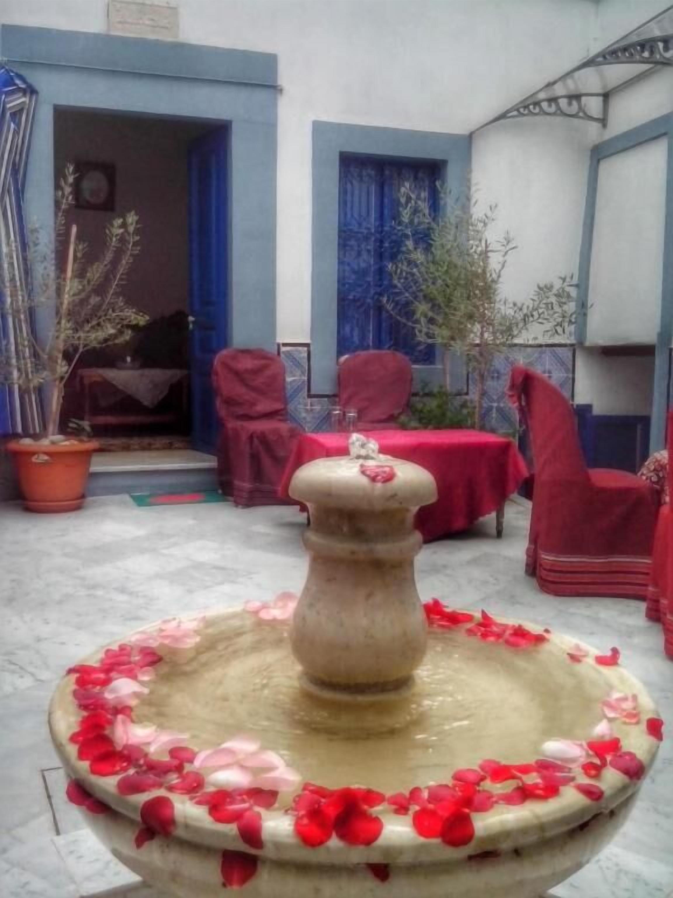 Un patio con una fuente central rodeada de sillas rojas y una mesa, plantas en macetas y ventanas con marcos azules.