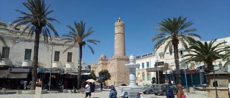 A square with a tall minaret, palm trees, and a fountain.
