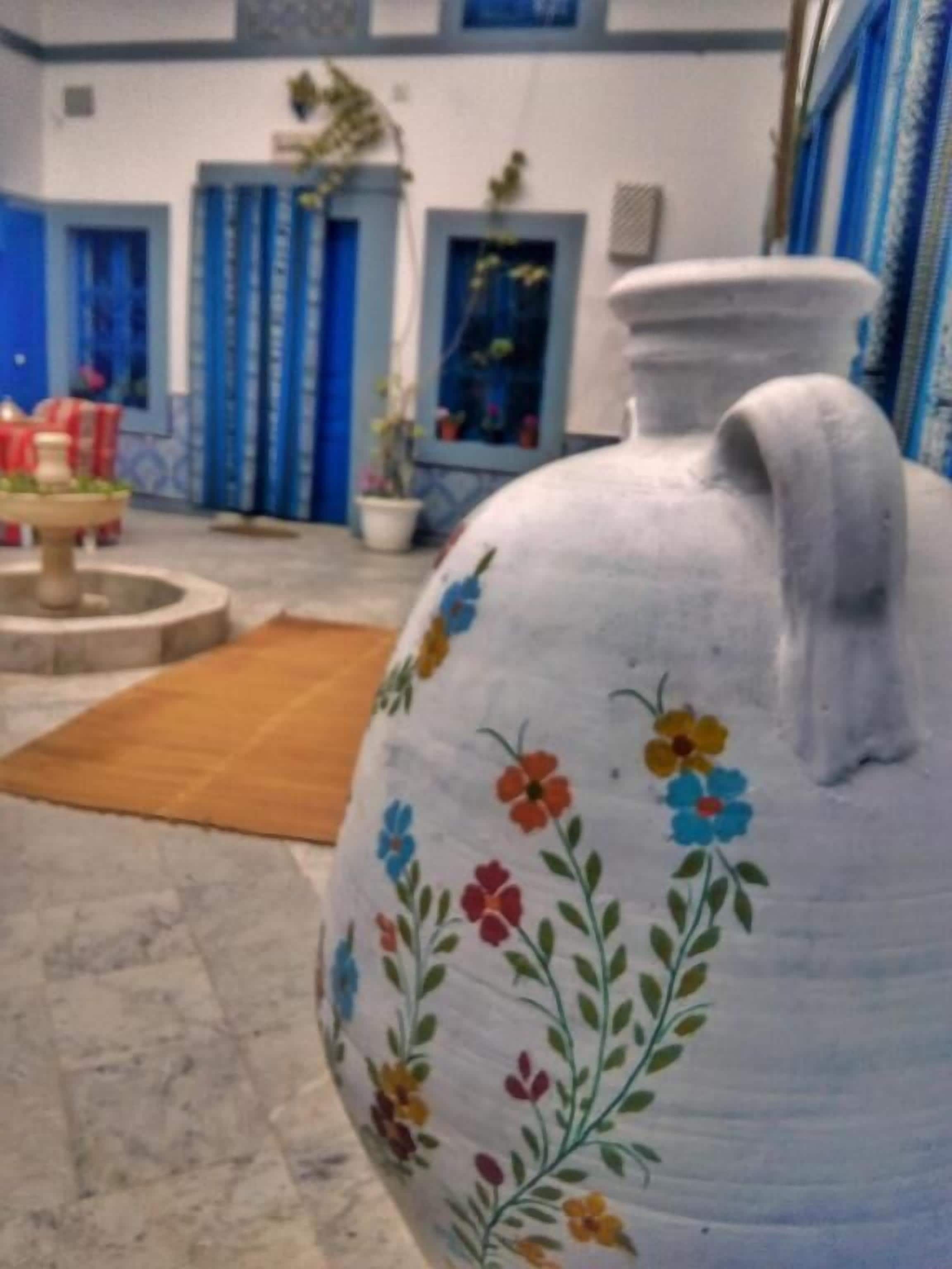 Un grand vase blanc en céramique, orné de motifs floraux colorés, est posé sur un sol carrelé dans une pièce aux portes bleues, où se trouve également une petite fontaine.