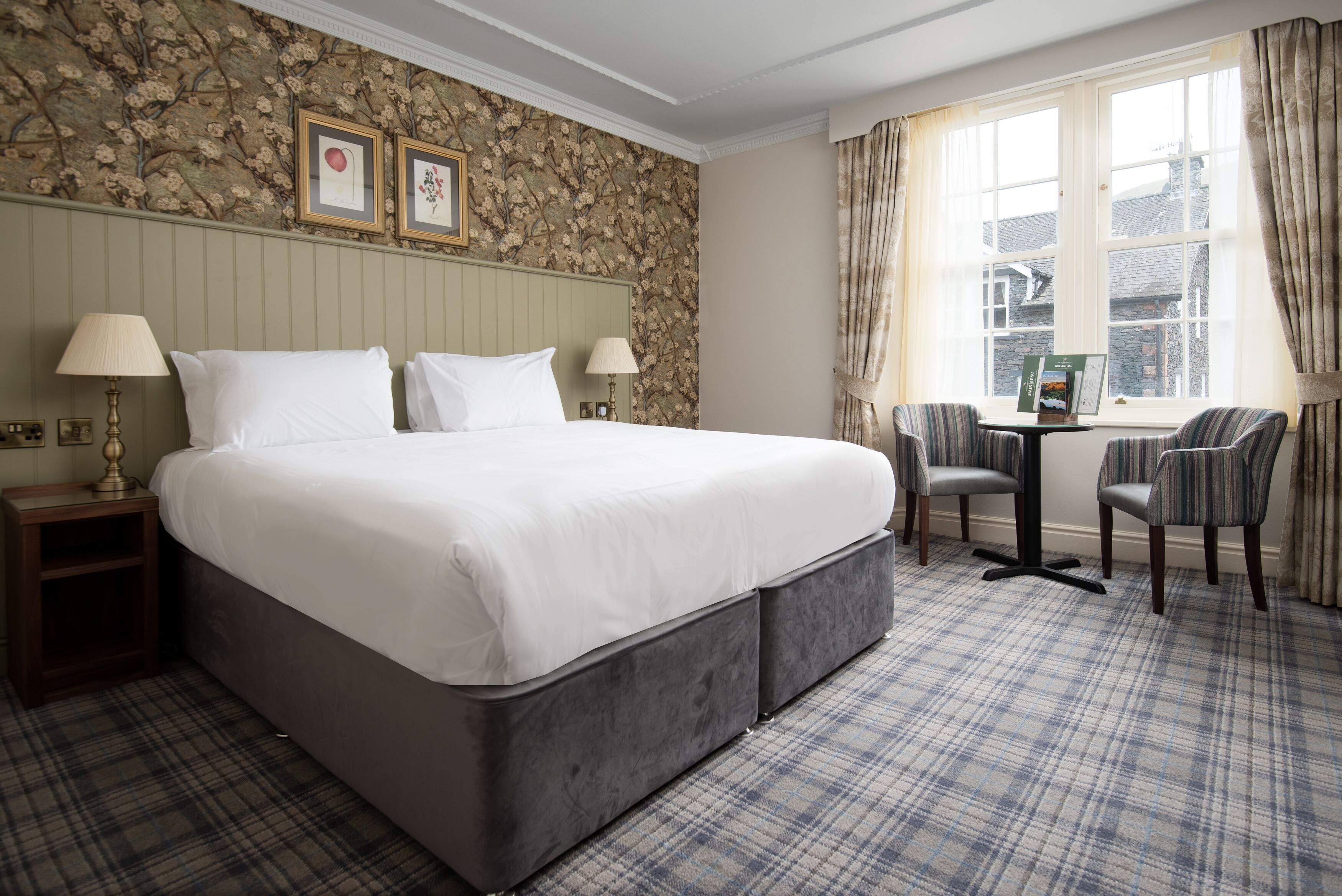 A hotel room with a large bed, two bedside tables with lamps, a small table with a book, two chairs, and a window with curtains.