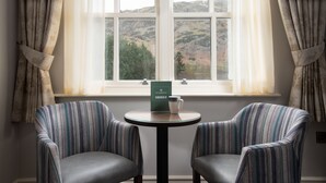 A room with two striped armchairs, a small round table with a menu, and a window with a view of hills. - The Ullswater Inn- The Inn Collection Group (Penrith)