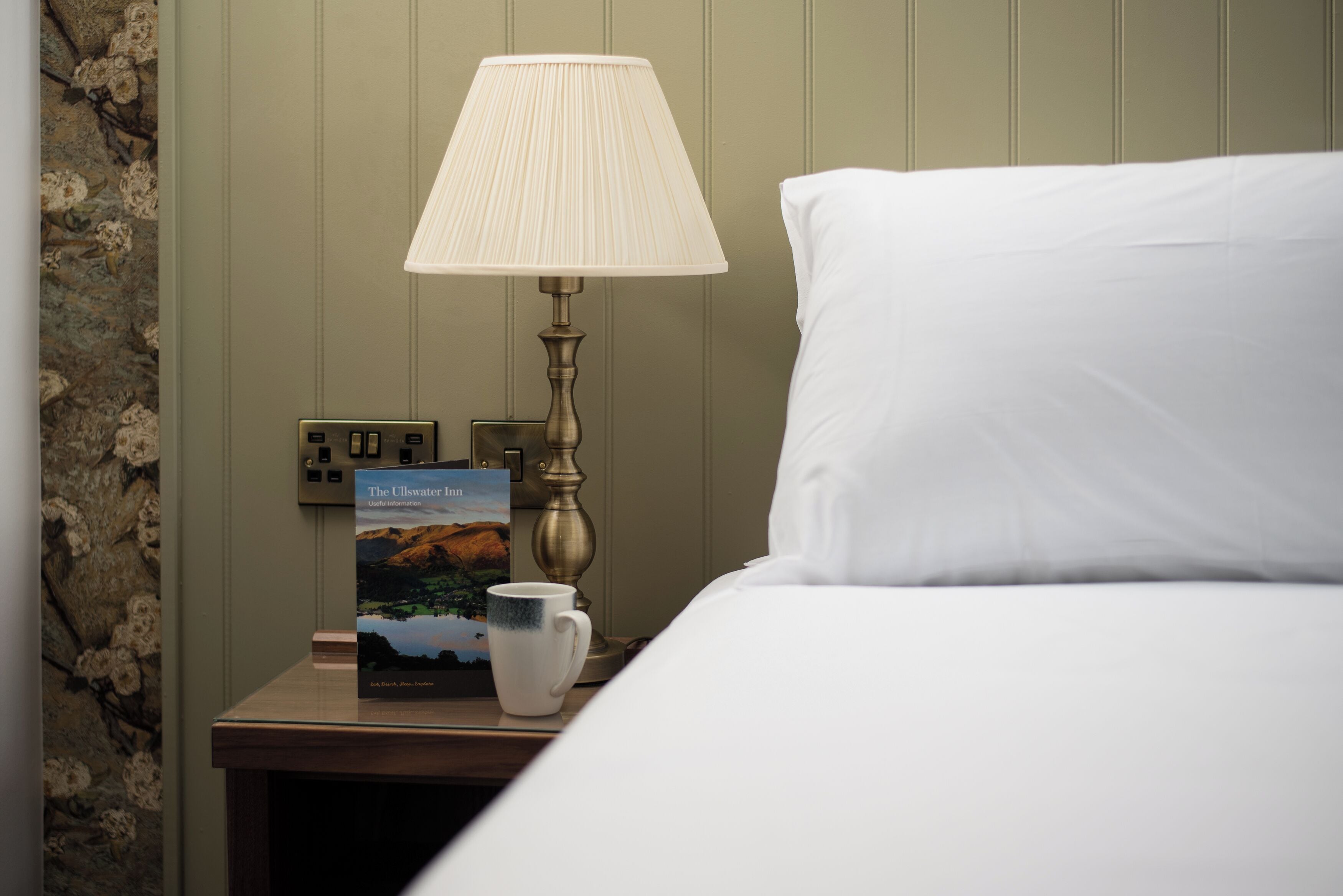 A neatly made bed with a white pillow and a white pillowcase. A bedside table holds a lamp, a book titled "The Ulster Inn," and a white mug. The wall behind the bed is painted in a light green color.