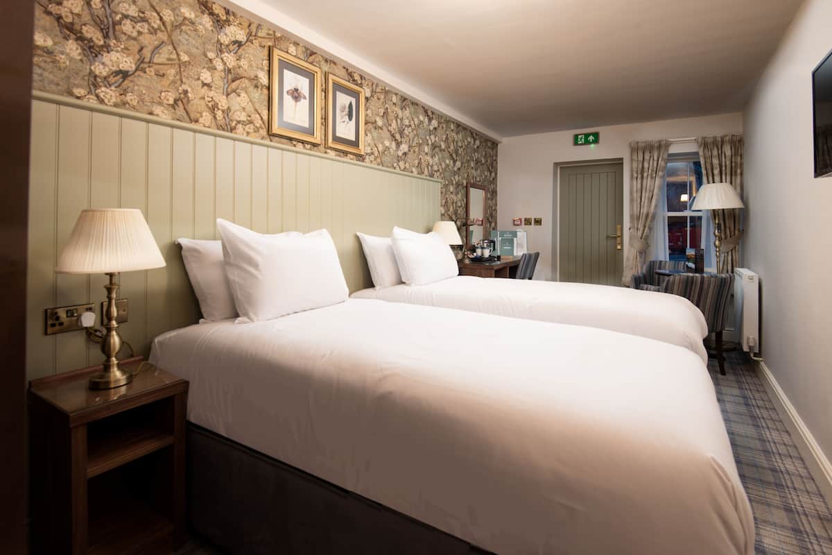 Habitación de hotel con dos camas, una mesita de noche de madera con una lámpara, un escritorio con una silla, un radiador y un papel tapiz con estampado floral.
