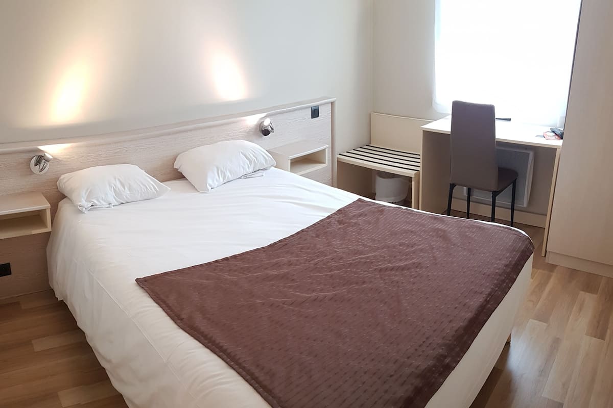 Un lit bien fait, avec une literie blanche et un plaid marron, un bureau avec une chaise, et une fenêtre laissant entrer la lumière naturelle.
