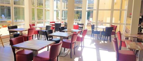 A spacious indoor area with multiple tables and chairs arranged for dining. Large windows allow natural light to fill the space.