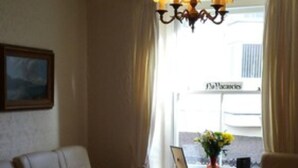 A living room with a sofa, a coffee table, and a chandelier. There is a window with curtains and a sign outside. - Myrtle House Hotel (Tenby)