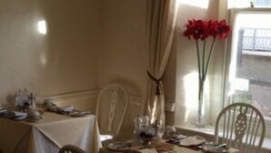 A dining area with a table set for two, chairs, a vase with red flowers, and a window with curtains. - Myrtle House Hotel (Tenby)