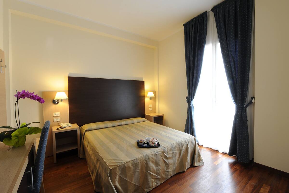 A hotel room with a bed, bedside lamps, a small table with a teapot and cups, a potted plant, and a window with curtains.