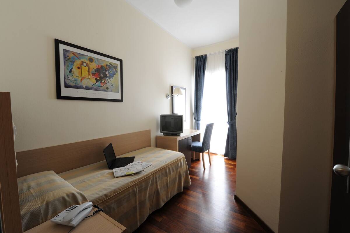A hotel room with a bed, a desk with a chair, a television, a framed picture on the wall, and a door.