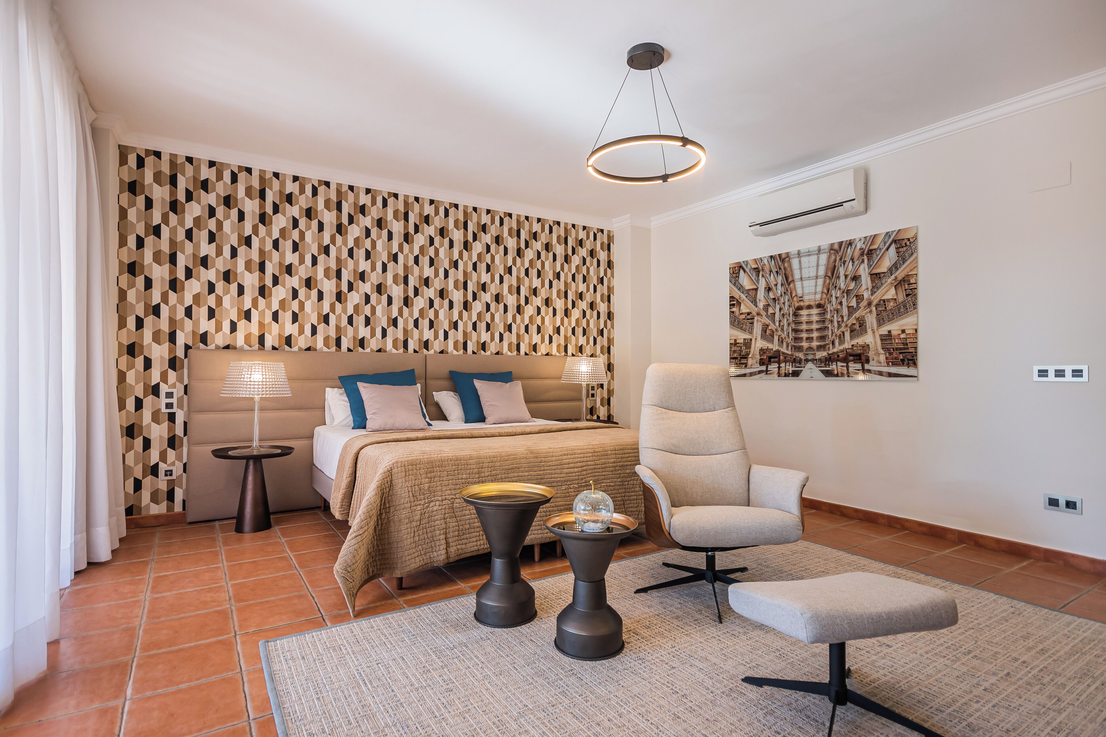 Un dormitorio moderno con una pared de diseño geométrico, una cama grande con ropa de cama beige y azul, un sillón blanco, una mesita de noche con lámpara y un amplio ventanal con vistas a una estructura arquitectónica.