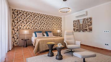Un dormitorio moderno con una pared de diseño geométrico, una cama grande con ropa de cama beige y azul, un sillón blanco, una mesita de noche con lámpara y un amplio ventanal con vistas a una estructura arquitectónica.