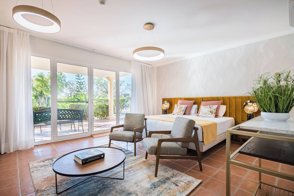 Une chambre moderne avec un grand lit, deux fauteuils, une petite table et une vue sur l’extérieur grâce à de grandes portes coulissantes.