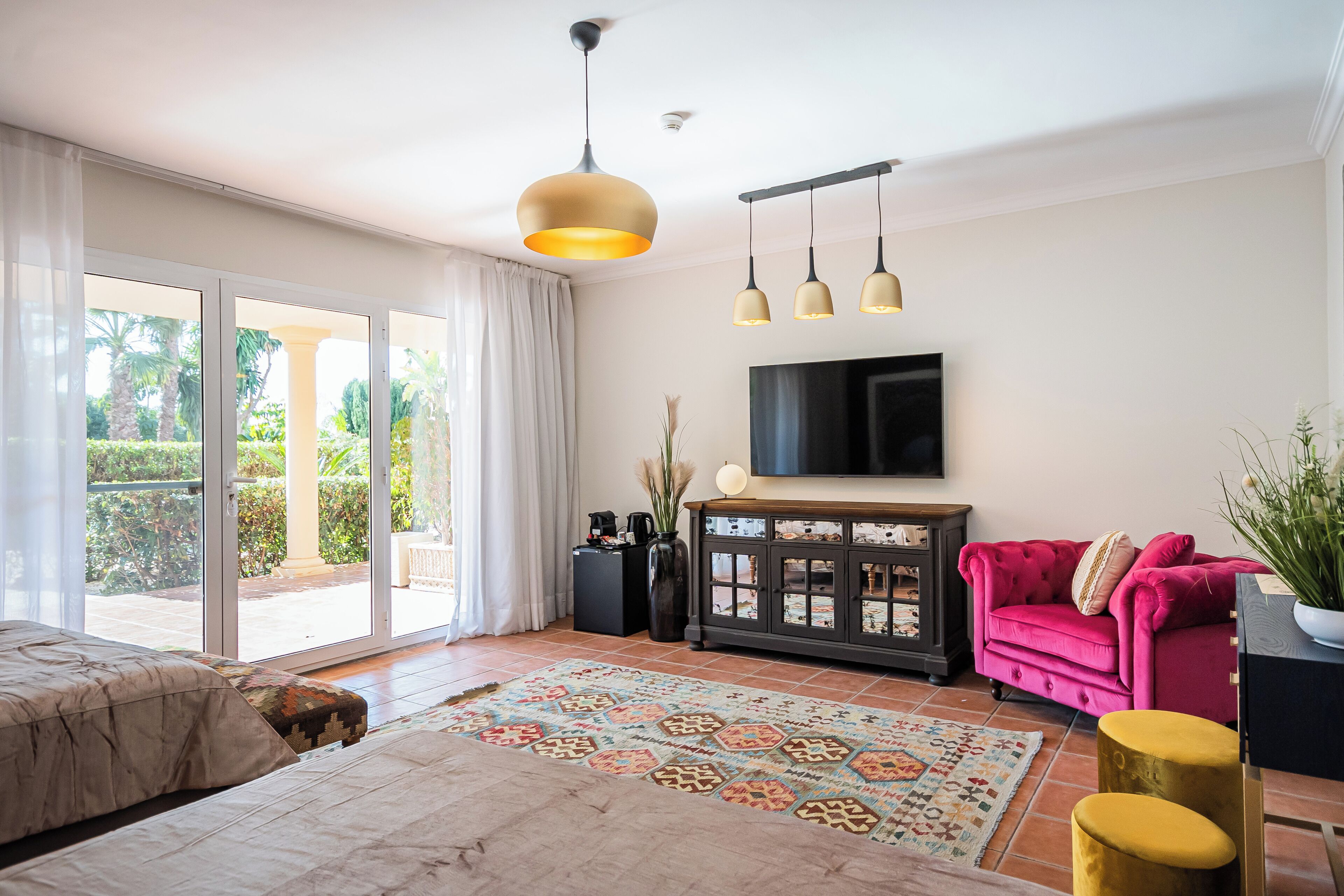 Un salón amplio con un gran ventanal corredera que da a un patio, un sofá rosa, un televisor montado en la pared y una alfombra estampada.