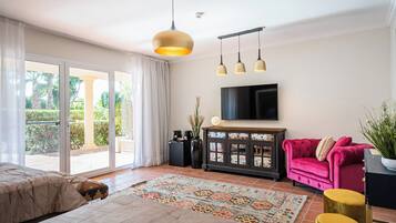 Un salón amplio con un gran ventanal corredera que da a un patio, un sofá rosa, un televisor montado en la pared y una alfombra estampada.