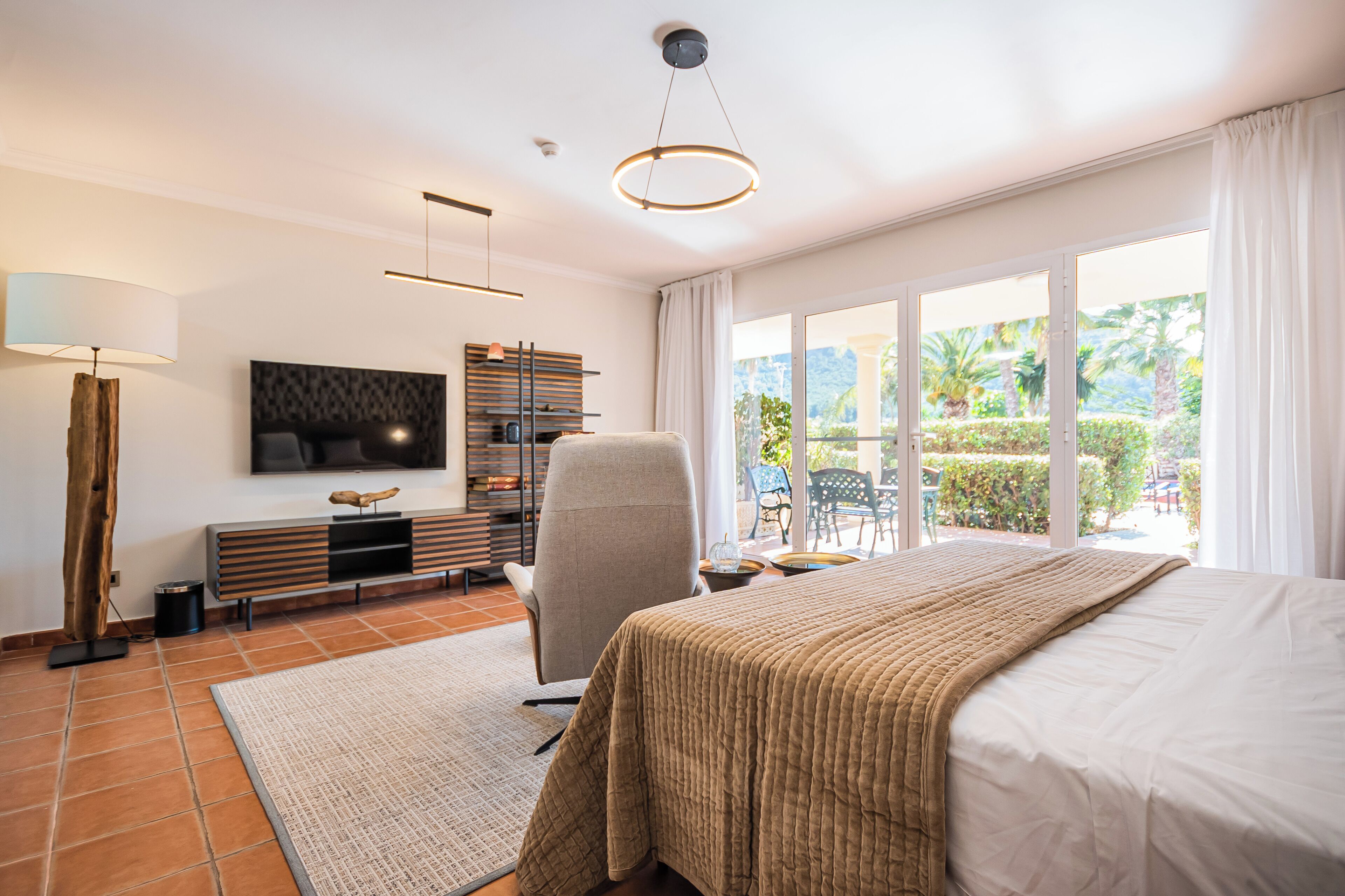 A modern bedroom with a large bed, a TV mounted on the wall, a chair, a desk, and a view of the outdoors.