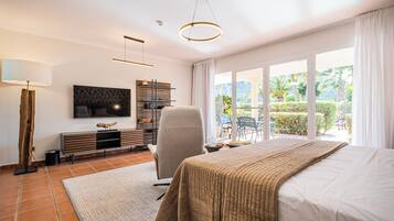 A modern bedroom with a large bed, a TV mounted on the wall, a chair, a desk, and a view of the outdoors.