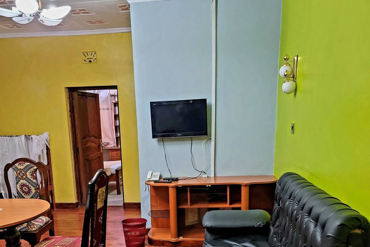A living room with a black leather sofa, wooden dining table, and chairs. A flat-screen TV is mounted on the wall.